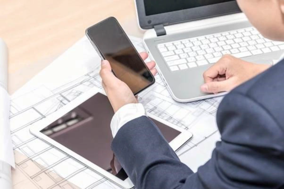 professional reviewing commercial property documents and architectural plans across laptop tablet and phone representing UK commercial mortgage underwriting stage